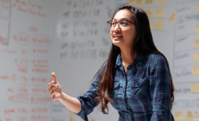 A person standing and speaking in front of a white board with data on it